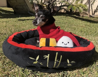 Pode incluir: Uma cama para cães preta e vermelha em forma de rolo de sushi com um cão preto e branco vestindo um suéter vermelho dentro. A cama tem um design de bambu verde no lado.