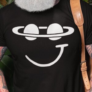 Peut inclure: T-shirt noir avec un motif blanc représentant un visage souriant avec des yeux en forme de planètes et une bouche incurvée. Le motif est centré sur le t-shirt. La personne porte un jean bleu et a des tatouages sur les bras.
