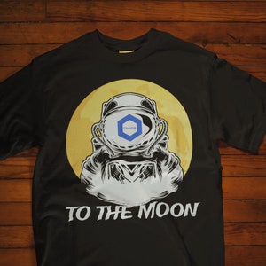 May include: Black t-shirt with a graphic of an astronaut in front of a yellow moon. The astronaut's helmet has a blue and white logo. The text "TO THE MOON" is printed below the graphic.
