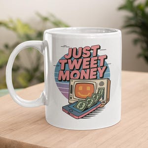 May include: White ceramic mug with the text "JUST TWEET MONEY" in pink and blue. The mug features a retro design with a smiling television, a smartphone, and dollar bills. The mug is on a wooden surface.