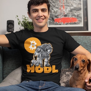 May include: A black t-shirt featuring a yellow Bitcoin logo and a cartoon character holding a Bitcoin. The text "HODL" is printed in yellow below the character. A person is wearing the shirt and sitting on a sofa with a brown dachshund.
