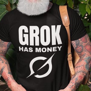 May include: Black t-shirt with the text "GROK HAS MONEY" in white, along with a stylized symbol. The shirt is worn by a person with a white beard and tattoos on their arms.