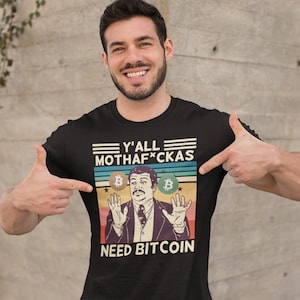 May include: A black t-shirt with a retro-style graphic featuring a man with a surprised expression holding two gold Bitcoin coins. The text on the shirt reads "Y'all Mothaf*ckas Need Bitcoin".