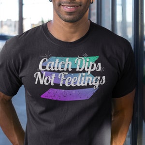 Op de afbeelding: Een zwart T-shirt met een grafische print die "Catch Dips Not Feelings" zegt in een retro stijl met een kleurverloop van groen, paars en blauw.