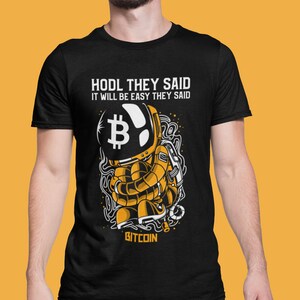 May include: Black t-shirt featuring a cartoon astronaut wearing a helmet with a Bitcoin symbol. The text "HODL THEY SAID IT WILL BE EASY THEY SAID BITCOIN" is printed on the shirt.