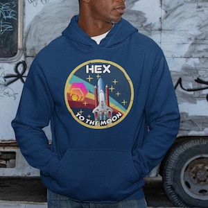 Puede incluir: Una sudadera con capucha azul marino con un gráfico de transbordador espacial de estilo retro y el texto "HEX TO THE MOON" en un círculo amarillo.