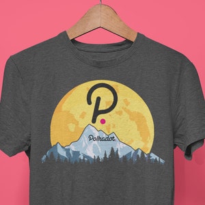 Op de afbeelding: Donkergrijs t-shirt met een afbeelding van een gele maan, bergen en bomen. Het Polkadot-logo staat boven de bergen. Het shirt hangt aan een houten hanger tegen een roze achtergrond.