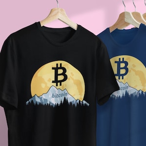 May include: Black and navy blue t-shirts with a Bitcoin graphic. The design features a Bitcoin symbol over a yellow moon, mountains, and the word "bitcoin". The shirts are hanging on a white rail.