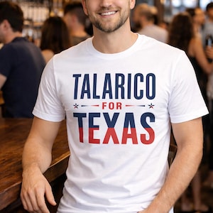 May include: White t-shirt with the text "TALARICO FOR TEXAS" in navy blue and red. The shirt is worn by a person in a bar setting.