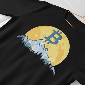 Könnte beinhalten: Schwarzes Sweatshirt mit einem gelben Mond und einem blauen Bitcoin-Symbol. Der Mond wird teilweise von einer Bergkette mit dem Wort "Bitcoin" verdeckt.