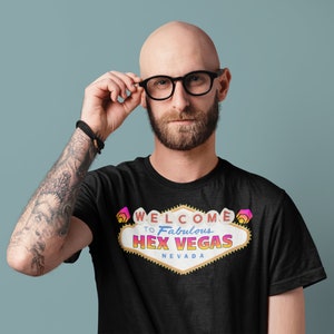 Puede incluir: Una camiseta negra con un gráfico de un letrero de neón que dice "Bienvenido al fabuloso Hex Vegas Nevada".