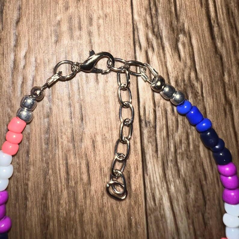 K&ouml;nnte beinhalten: Eine farbenfrohe Perlenkette mit Silberverschluss und Verl&auml;ngerungskette. Die Perlen sind in den Farbt&ouml;nen Rosa, Wei&szlig;, Lila, Blau und Silber gehalten. Die Halskette wird auf einer Holzoberfl&auml;che pr&auml;sentiert.