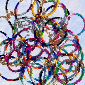 May include: A collection of colorful beaded bracelets in various shades, including pink, blue, green, yellow, and orange. The bracelets have gold and silver chain accents and clasps. The beads are small and round, creating a vibrant and playful aesthetic.