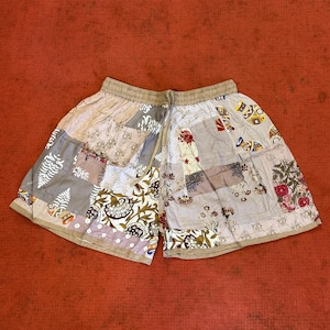 May include: Patchwork shorts with a drawstring waist. The shorts feature a variety of patterned fabric squares in shades of beige, grey, and floral prints. The shorts are laid flat on a red surface.
