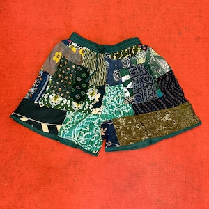 Op de afbeelding: Patchwork shorts in verschillende tinten groen, blauw en goud. De shorts hebben een mix van stoffen met patronen, met een groene tailleband en bies. Het kledingstuk ligt plat op een rode ondergrond.
