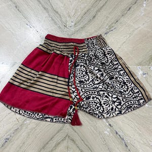 May include: A pair of patterned shorts with a red waistband and drawstring. One side features a red and tan striped design, while the other has a black and white floral pattern. The shorts have a red tassel on the drawstring.