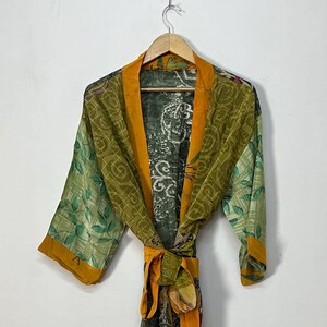 May include: A long-sleeved robe featuring a patchwork design with various green, olive, and mustard yellow patterns. The garment includes a solid mustard yellow collar and a matching tie belt at the waist, hanging from a wooden hanger.