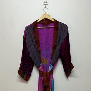 May include: A colorful, patchwork robe with a belt, hanging on a wooden hanger. The robe features a mix of patterns and colors, including purple, pink, blue, and brown. The sleeves and trim are a darker shade of purple and brown.
