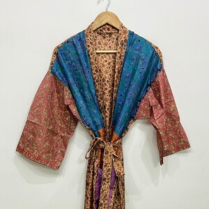 May include: A colorful, patterned robe with a tie closure. The robe features a mix of floral and paisley designs in shades of teal, coral, and tan. The robe is hanging on a wooden hanger against a white background.