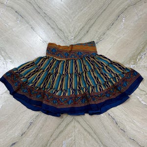 Puede incluir: Falda escalonada con estampado. La falda presenta rayas verticales alternas en verde azulado y dorado, con un borde y una cintura azul oscuro. La cintura tiene un diseño estampado en tonos marrones, naranjas y azules. La falda mide unos 60 cm de largo.