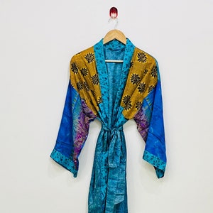 May include: A long, flowing robe in shades of teal, blue, and gold, hanging on a wooden hanger. The robe features a floral pattern on the shoulders and sleeves, with a matching tie belt. The fabric appears to be lightweight and silky.
