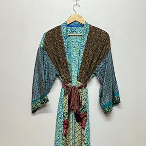 May include: A long-length robe featuring a patchwork design with various floral and geometric patterns in shades of teal, light blue, dark brown, and cream. The garment includes wide sleeves, a front opening, and a matching fabric tie belt at the waist.