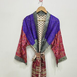May include: A colorful patchwork robe with a tie belt. The robe features a mix of patterns, including paisley, stripes, and floral designs, in shades of purple, red, and green. The robe is hanging on a wooden hanger.