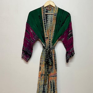 May include: A tie-dye kimono robe in shades of green, purple, black, and orange hangs on a wooden hanger. The robe features a tie-dye pattern and a matching belt. The sleeves are a solid green and purple.