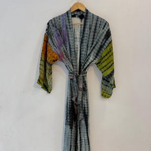 May include: A long-sleeved kimono robe featuring a tie-dye pattern in shades of grey, black, yellow, and purple. The garment includes a matching fabric tie belt at the waist, an open front design, and wide sleeves, all displayed on a wooden hanger.