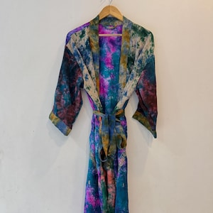 May include: A long-sleeved robe featuring a vibrant tie-dye pattern in shades of deep blue, magenta, teal, and olive green. The garment includes a matching tie belt at the waist, wide sleeves, and a shawl collar, all displayed on a wooden hanger.