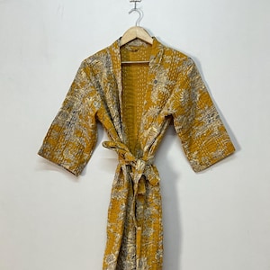 May include: Mustard yellow robe with a floral pattern in gray and cream. The robe has a tie belt and three-quarter sleeves. The garment is hanging on a wooden hanger against a white wall.