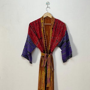 Può includere: Una vestaglia a maniche lunghe con motivo tie-dye nei toni del rosso, viola e marrone. Il capo presenta una cintura in tessuto abbinata in vita, un design aperto sul davanti e maniche larghe, appeso a una gruccia di legno.