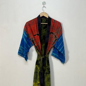May include: A tie-dye robe with a vibrant color scheme. The top features red and blue sections, while the body transitions from green to black. A matching green tie belt cinches the waist. The robe is hanging on a wooden hanger.