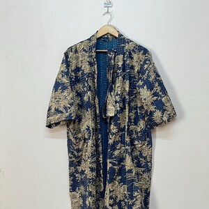 May include: A long-sleeved robe in a deep blue hue featuring a beige floral pattern throughout. The garment is constructed with visible white kantha stitching and includes a matching fabric belt, hanging on a wooden hanger against a plain wall.