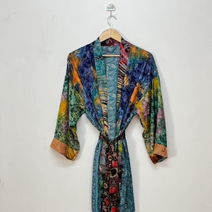 Könnte beinhalten: Ein langärmeliger Kimono-Bademantel aus einem Patchwork aus leuchtend bunten Stoffen mit verschiedenen Mustern, darunter Paisley, Blumen- und abstrakte Designs. Das Kleidungsstück hat einen passenden Bindegürtel und hängt an einem Holzkleiderbügel.