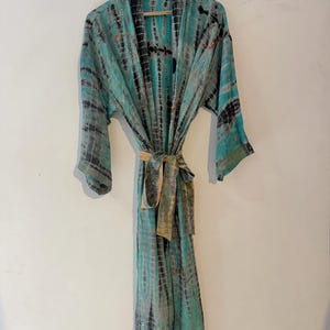 Può includere: Una vestaglia a maniche lunghe con motivo tie-dye nei toni del verde acqua, nero e grigio. Il capo presenta una cintura in tessuto abbinata in vita e sottili decorazioni con perline metalliche lungo i risvolti anteriori e le spalle.