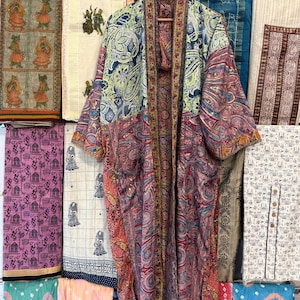 May include: A long-sleeved kimono-style robe featuring a patchwork design of various paisley and floral patterns in shades of blue, red, yellow, and green. The garment has an open front with a contrasting patterned trim along the collar and edges.