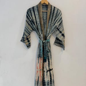 Könnte beinhalten: Ein langärmeliger Morgenmantel mit einem Batikmuster in den Farben Anthrazit, gedämpftem Blau und zartem Pfirsich. Das Kleidungsstück hat eine offene Vorderseite mit einem passenden Bindegürtel und einen einfarbigen Kragen in Beige.