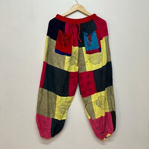 May include: Loose-fitting patchwork trousers featuring an elasticized waistband with a red drawstring. The pants are constructed from rectangular panels in black, red, yellow, and tan, accented with black floral and paisley print patterns.