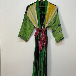 May include: A long, tie-dye robe in shades of green, yellow, and black, with a matching belt. The robe is hanging on a wooden hanger against a white background. The robe's design features vertical stripes and a colorful pattern.