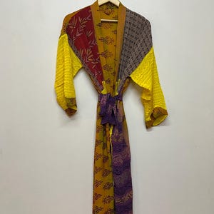 May include: A patterned robe with long sleeves, hanging on a wooden hanger. The robe features a mix of yellow, red, and purple fabrics with various designs. A matching belt is tied around the waist.