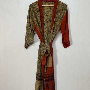 May include: A long-sleeved robe featuring a mustard yellow and black patterned fabric with a repeating circular design. The garment includes solid burnt orange trim along the front opening, sleeves, and a matching tie belt at the waist.