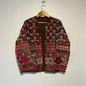 Può includere: Giacca marrone senza chiusura con ricami intricati. La giacca, appesa a una gruccia di legno, presenta un motivo dettagliato in rosso, rosa, blu e verde. Lo sfondo è una parete bianca.