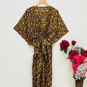 May include: A black kaftan dress featuring a repeating golden yellow floral pattern, wide kimono-style sleeves, and a cinched waist detail. The garment is displayed on a wooden hanger against a plain background, showcasing a V-neckline.