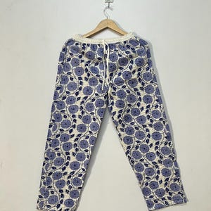Peut inclure: Pantalon crème avec un motif floral bleu. Le pantalon est suspendu à un cintre en bois contre un mur blanc. Le motif floral présente de grandes fleurs et feuilles bleues stylisées. Le pantalon a une taille à cordon.