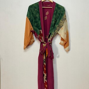 May include: A vibrant, patterned robe with a belt. The robe displays a mix of colours, including green, orange, and magenta. The robe is hanging on a wooden hanger against a white wall. The robe has a floral pattern on the lower section.