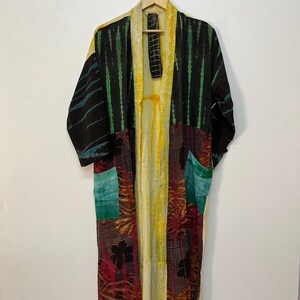 May include: A long, patchwork kimono robe with a variety of patterns and colors. The robe features black fabric with green vertical stripes, yellow and cream-colored panels, and patches of red, pink, and teal. It hangs on a wooden hanger.