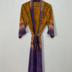 Op de afbeelding: Een lange, vloeiende kimono met een bloemenpatroon in tinten geel, oranje en paars. De kimono heeft een bijpassende ceintuur en hangt aan een houten hanger. De mouwen zijn driekwart en de stof lijkt lichtgewicht.