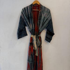 May include: A long-sleeved robe featuring a tie-dye pattern in shades of deep red, charcoal gray, and muted blue. The garment has an open front design with a matching tie-dye fabric belt cinched at the waist, hanging from a wooden hanger.