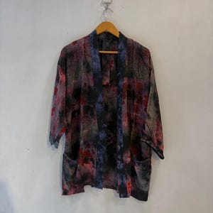 May include: A lightweight open-front kimono jacket featuring a dark, moody floral pattern in shades of black, deep red, and muted purple. The garment has three-quarter length sleeves, two front patch pockets, and a contrasting blue patterned trim.